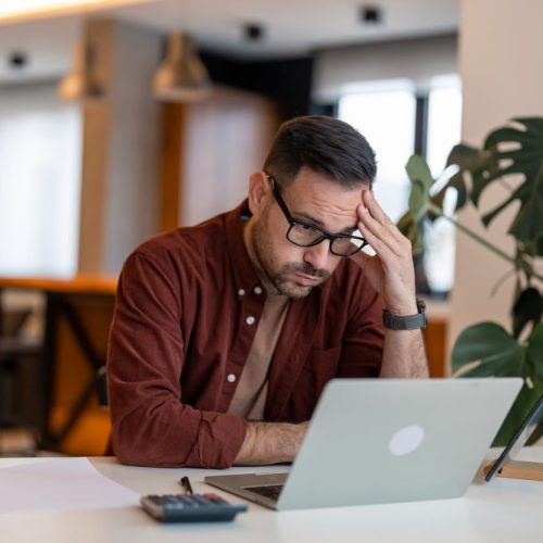 Contemplating businessman in the home office feeling frustrated. Full concentration on work. Man working on laptop, counting profit while sitting at the desk at home.